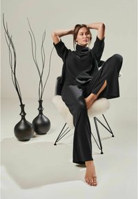 Schwarzer, gerippter Pullover mit hohem Kragen, kombiniert mit lockeren schwarzen Hosen, sitzt auf einem Stuhl mit schwarzen Vasen und dekorativen Ästen im Hintergrund.