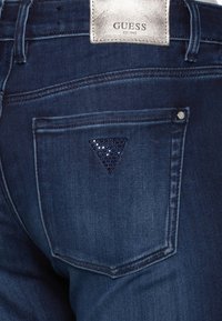 Jeans de mezclilla de lavado oscuro con una etiqueta plateada de "Guess". Presenta un bolsillo trasero con un diseño de triángulo de rhinestone azul y detalles de costura.