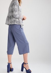 Ljust grå spetsblus med peplum nederkant, tillsammans med lösa, korta blå byxor. Modellen bär blå högklackade sandaler. Släta strukturer, mjuka nyanser.