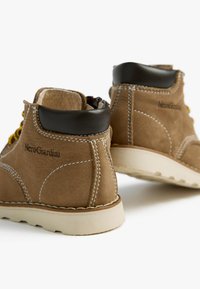 Braune Wildleder-Stiefeletten mit einer tanfarbenen Gummisohle, schwarzen Lederdetails, gelben Schnürsenkeln und kontrastierenden weißen Nähten. Logo an der Seite sichtbar.