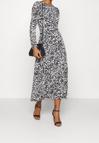 Zwart-wit gestreepte jurk met lange mouwen en een uitlopende rok, voorzien van knoopdetails, gecombineerd met een zwarte clutch en sandalen met hakken.