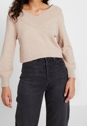 Femme portant un pull beige à col en V avec des manches bouffantes, associé à un jean noir taille haute, sur un fond uni.
