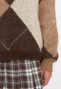 Close-up van een hand aan de zijkant die een kleurrijke fuzzy trui draagt in bruin, beige, crème en een bruine geruite rok.