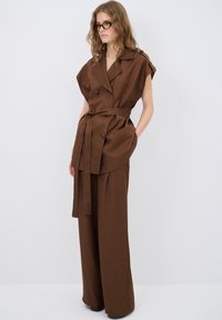 Gilet marrone su misura con maniche larghe, doppi bottoni e una cintura, abbinato a pantaloni larghi coordinati. Tessuto testurizzato, design minimalista.
