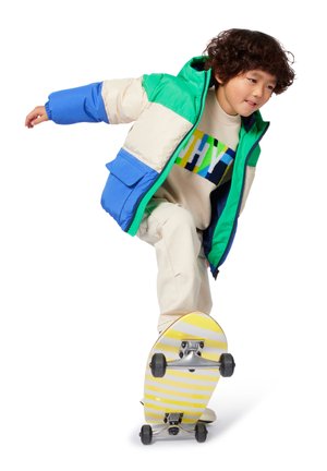 Bunte, gepolsterte Jacke mit grünen, blauen und cremefarbenen Panels, kombiniert mit einem beigen Pullover und cremefarbenen Hosen, auf einem gestreiften Skateboard fahrend.