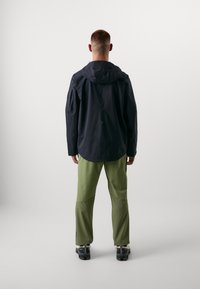 Veste imperméable noire avec capuche, portée par-dessus un pantalon vert clair. Présente un dos plus long, et les deux vêtements sont conçus pour des activités de plein air.