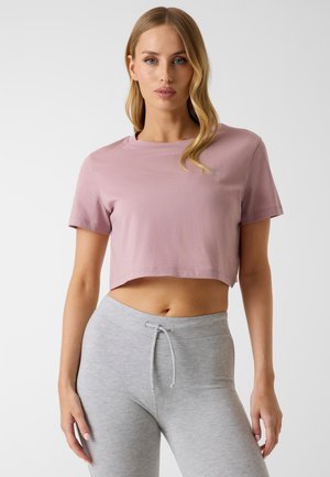 Frau mit langen blonden Haaren trägt ein hellrosa, bauchfreies T-Shirt und hellgraue Leggings mit Kordelzug, stehend vor weißem Hintergrund.