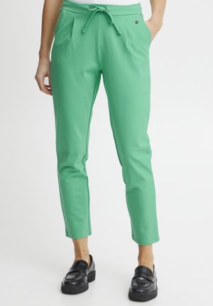 Pantalones de tela elástica verde con cintura ajustable, corte tapered y pliegues en la parte delantera. Presenta un pequeño detalle con logo.
