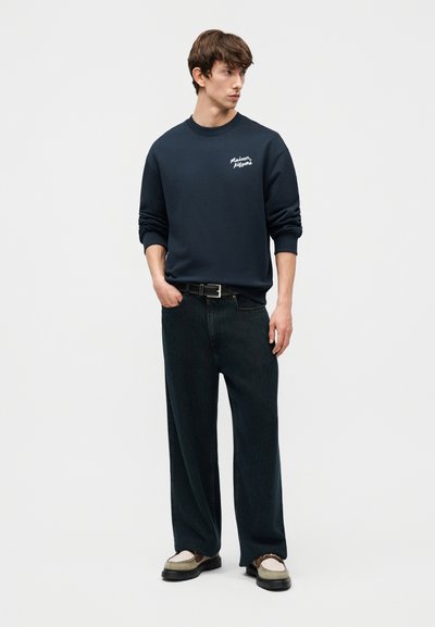 Άνδρας που φοράει μπλε ναυτικό φούτερ με το κείμενο «maison kitsune», μαύρο παντελόνι με φαρδιά πόδια, μαύρη ζώνη και μπεζ loafers, στέκεται με το ένα χέρι στην τσέπη.