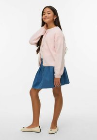 Šviesiai rožinis megztas kardiganas su sagomis, dėvimas ant balto viršaus ir mėlynos džinsinės sijono, derinamas su kreminės spalvos bateliais su sidabriniu akcentu.