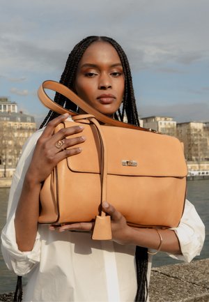 Vrouw met lang gevlochten haar houdt grote beige leren handtas vast buiten bij water en gebouwen, draagt wit overhemd en gouden sieraden.