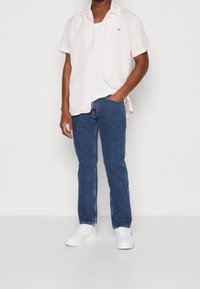 Světle růžová košile s krátkými rukávy, bílý tílko, modré džínové kalhoty a bílé tenisky, s malým logem na košili.