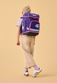 Dunkellila Rucksack mit pinkem Schuppenmuster, mehreren Fächern und Abzeichen. Kind trägt beige Hosen und weiße Schuhe mit marineblauen Streifen.