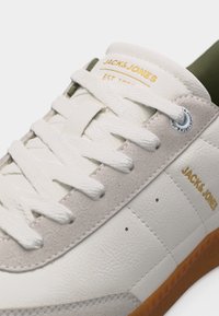 Λευκό και μπεζ αθλητικό παπούτσι Jack & Jones με λευκά κορδόνια, χρυσό λογότυπο στη γλώσσα και στο πλάι, και καφέ λαστιχένια σόλα, κοντινή λήψη.