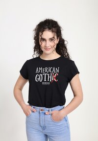 Mladá žena s kudrnatými vlasy, oblečená v černém tričku "American GothX Wednesday" a světlemodrých džínech, s rukama v kapsách.