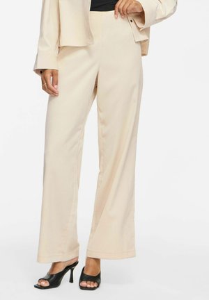 Femme portant un pantalon beige à jambes larges et une veste assortie, un haut noir et des chaussures à talons ouverts noires, debout sur un fond blanc.