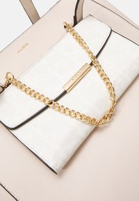 Beige kabelka s texturovaným bílým klopou, zdobená zlatým řetízkem a zlatou logovou destičkou. Hladká kůže.