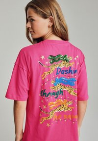 Rosa Baumwoll-T-Shirt mit gestickten Leoparden, die bunte Schals tragen, goldenen Sternen und dem Text "Durch den Schnee flitzen" in fetten Buchstaben.