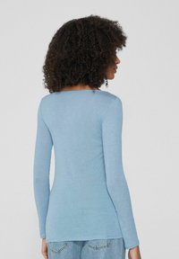 Hellblaue Langarmbluse mit rundem Ausschnitt und geradem Schnitt. Weicher Stoff, taillierte Passform und seitliche Schlitze am Saum.