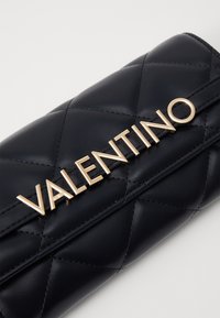 Borsa in pelle trapuntata blu navy con un prominente logo dorato "Valentino". Texture liscia con una forma rettangolare elegante e dettagli cuciti.
