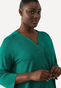 Teal långärmad klänning med V-ringning och subtila veck. Har en slät textur och en draperad design. Guld rektangulära örhängen.