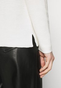 Biały sweter w prążki z bocznymi rozcięciami, noszony na czarnych skórzanych spodniach. Sweter ma okrągły dekolt i prążkowane mankiety.