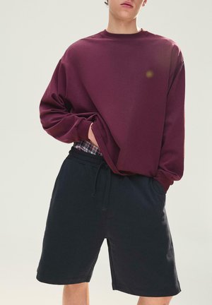 Persoană purtând un hanorac larg bordo și pantaloni scurți negri cu șnur, cu o mică vedere a chiloților în carouri la talie.