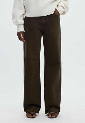 Personne portant un pantalon large marron foncé, un pull en tricot crème et des sandales noires, debout avec une main dans la poche.