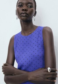 Sleeveless top in vibrant purple with black polka dots. Fabric is smooth with a round neckline. Model wears silver hoop earrings and a ring.