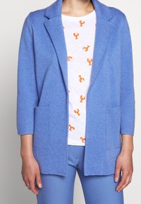 Ljusblå, texturerad kofta med notchad krage och sidofickor, bärs över en vit skjorta med orange hummermönster.