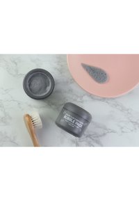 Un pot gris étiqueté "Pack de Bulles Carbonatées d'Argile Colorée" à côté d'une assiette rose contenant un échantillon d'argile grise, et un pinceau en bois avec des poils blancs.