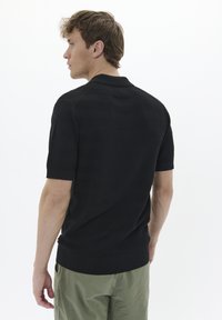 Črna polo majica s kratkimi rokavi, ki ima teksturiran vzorec, ravni ovratnik in rebrast rob, v kombinaciji s svetlo zelenimi hlačami.