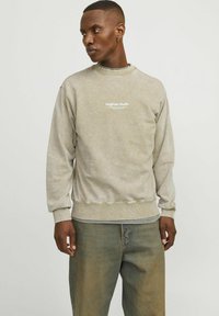 Sudadera de color beige claro con tejido texturizado, puños y dobladillo acanalados. Presenta el texto "Originals Studio" en blanco en el pecho. Corte casual.