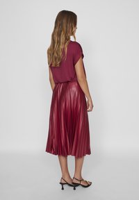 Bordeaux-farbener, plissierter Midirock mit glänzender Textur, kombiniert mit einem locker sitzenden, bordeauxfarbenen Oberteil und schwarzen Riemchenschuhen.