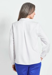 Weiße Bluse mit langen Ärmeln, die einen dezenten Rückenfalten sowie einen klassischen Kragen aufweist, kombiniert mit blauen Hosen für einen schönen Kontrast. Glatte Textur.