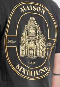 Zwart T-shirt met een gouden architecturale tekening van een gebouw, met de tekst "MAISON," "PARIS," en "SIXTH JUNE" duidelijk zichtbaar.