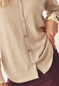 Cardigan in maglia beige con chiusura a bottoni frontale composta da cinque bottoni, vestibilità rilassata e orlo curvo. Tessuto morbido e texture evidente.