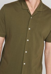 Kortärmad olivgrön skjorta med knappar, tillverkad av mjuk bomull, med krage och fem vita knappar fram.
