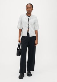 Γυναίκα που φοράει ανοιχτό γκρι ζακέτα με κοντά μανίκια πάνω από μαύρο τοπ και παντελόνι, κρατώντας μαύρη τσάντα, στέκεται μπροστά σε λευκό φόντο.