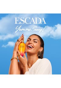 Glunderende vrouw met gouden sieraden houdt oranje en roze Escada-parfumfles vast tegen een felblauwe lucht met witte wolken.