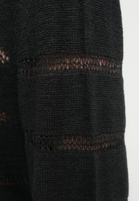 Μαύρο πλεκτό πουλόβερ με υφή ύφασματος που περιλαμβάνει χοντρές ενότητες και ανοιχτά σχέδια ύφανσης. Κοντά μανίκια με άνετη εφαρμογή.