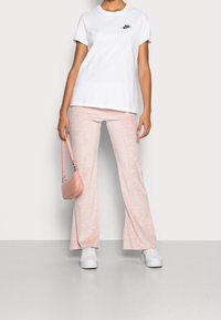 Person som bär en vit Nike t-shirt, ljusrosa mönstrade byxor, vita sneakers, och håller en liten rosa handväska framför en enfärgad bakgrund.