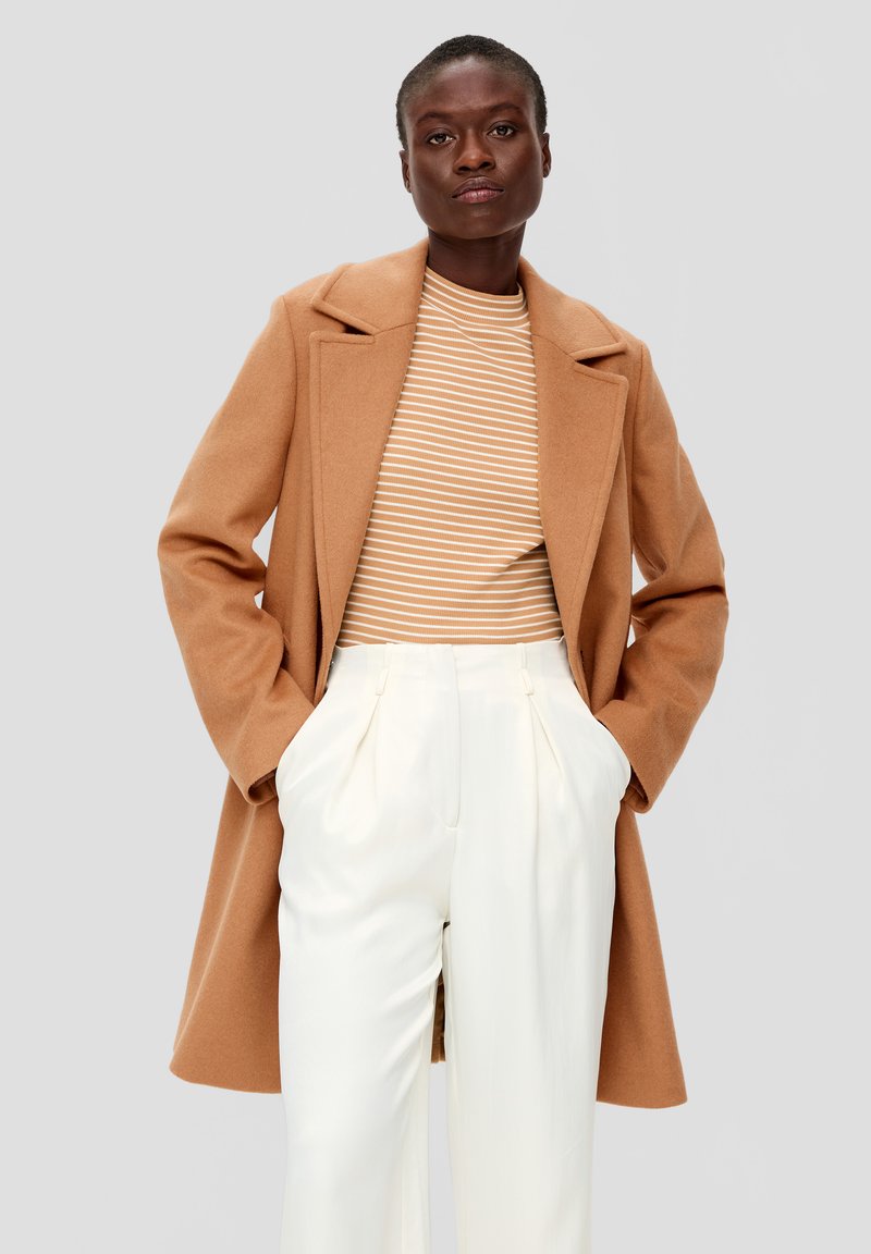 Bruin wollen jas met een gekartelde kraag over een beige en witte gestreepte top, gecombineerd met hoge witte broeken. Glad textuur.