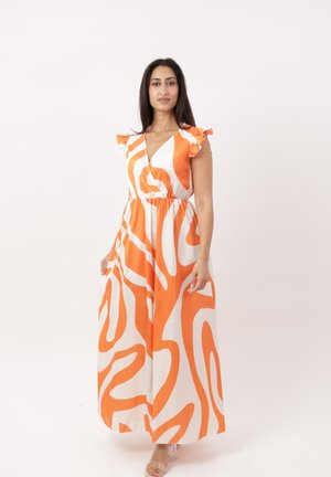 Mujer vestida con un maxi vestido sin mangas con estampado naranja y blanco y hombros con volantes, de pie contra un fondo claro y liso.