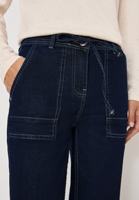Pantaloni in denim blu scuro con due tasche frontali, cuciture bianche a contrasto e chiusura con cordino in vita. Texture liscia.