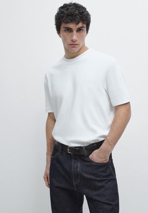 Giovane uomo con capelli ricci che indossa una maglietta bianca semplice a maniche corte infilata in jeans blu scuro con cintura nera, con una mano in tasca.