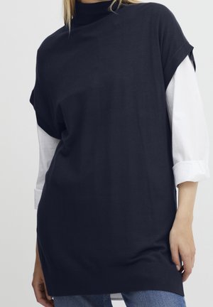 Vrouw die een zwarte mouwloze coltrui draagt over een wit overhemd met lange mouwen en een blauwe spijkerbroek, staand tegen een effen achtergrond.