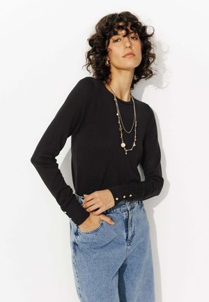 Haut noir à manches longues avec un col rond, ornements de boutons sur les poignets, associé à un jean en denim bleu clair taille haute.