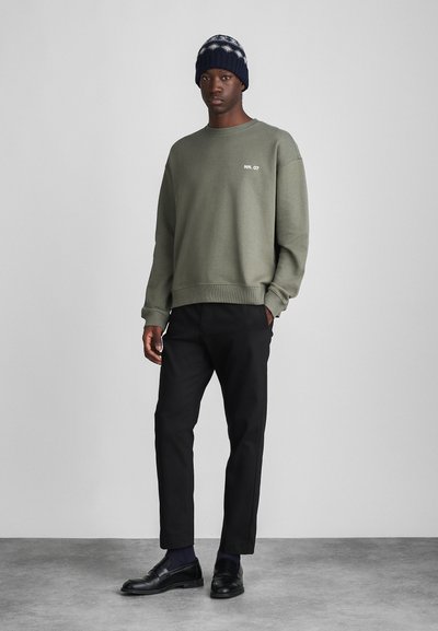 Olijfgroen sweatshirt met ribbels aan de manchetten, gecombineerd met zwarte rechte broek en zwarte instappers. Een donker gebreide beanie met patroon maakt de look compleet.