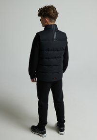 Gilet imbottito nero con colletto alto e cuciture orizzontali, abbinato a pantaloni neri e scarpe nere con dettagli bianchi.
